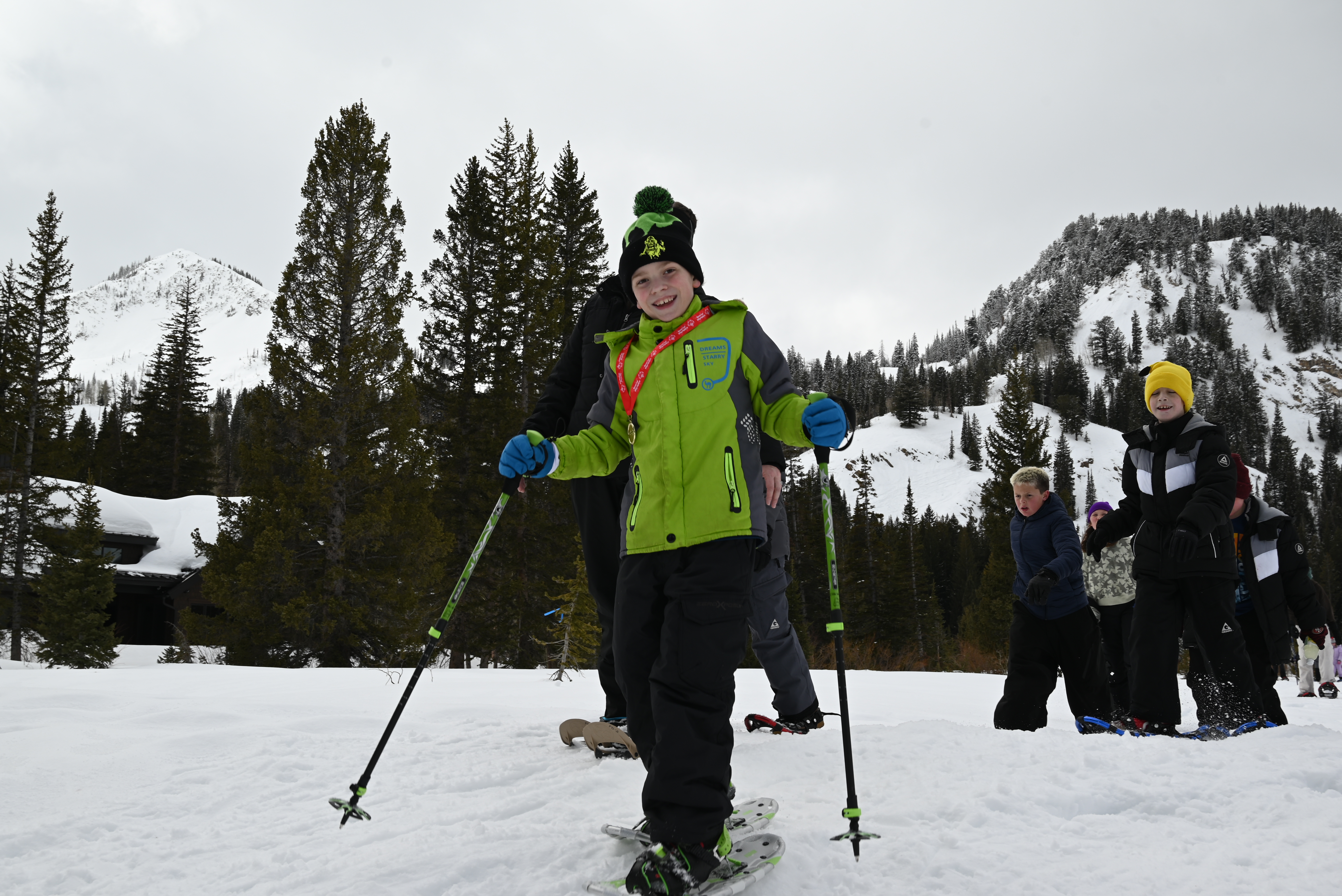 A boy in a green jacket snowshoeing with poles on the trail