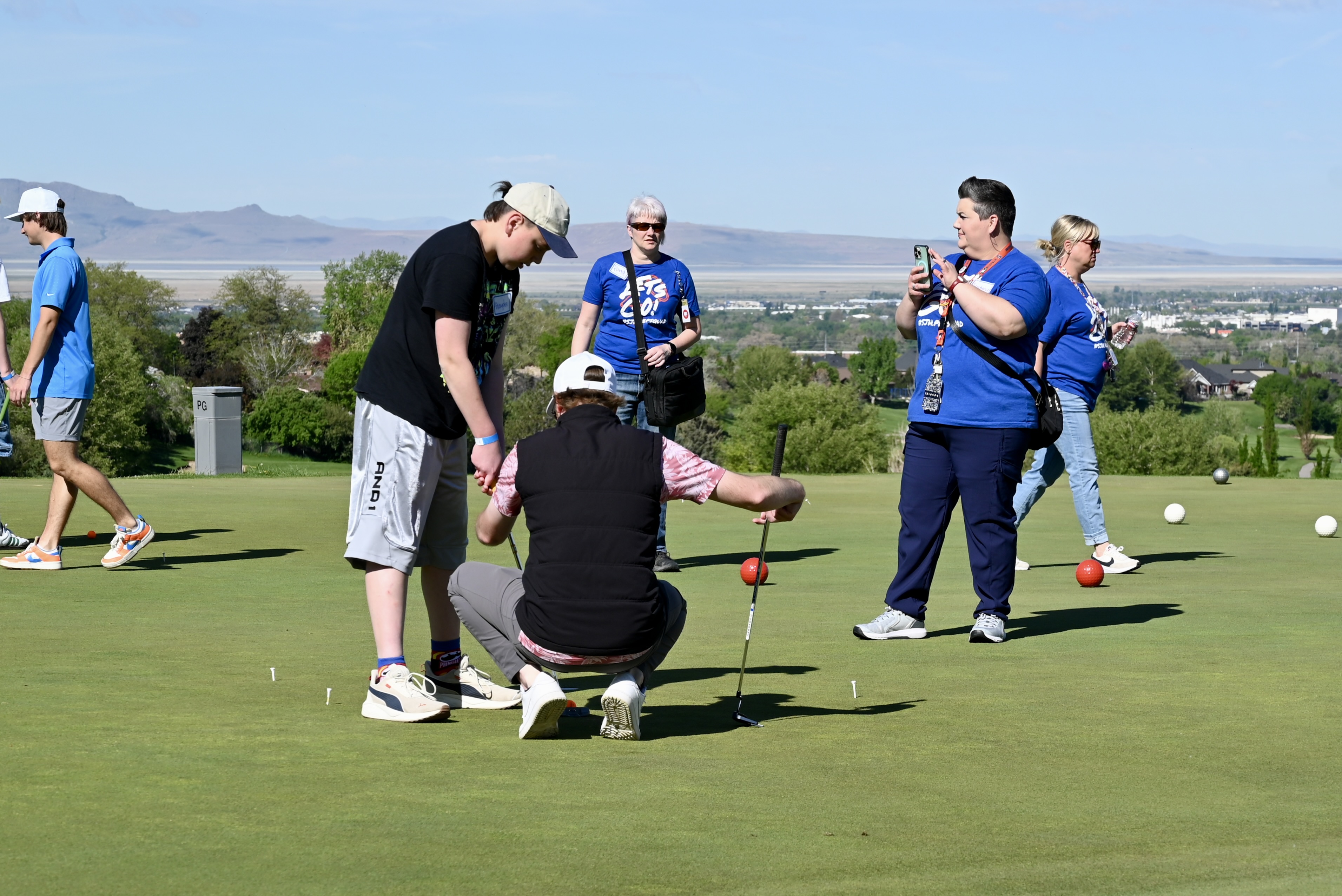 A coach works with students on putting while spectators watch with valley panorama behind them
